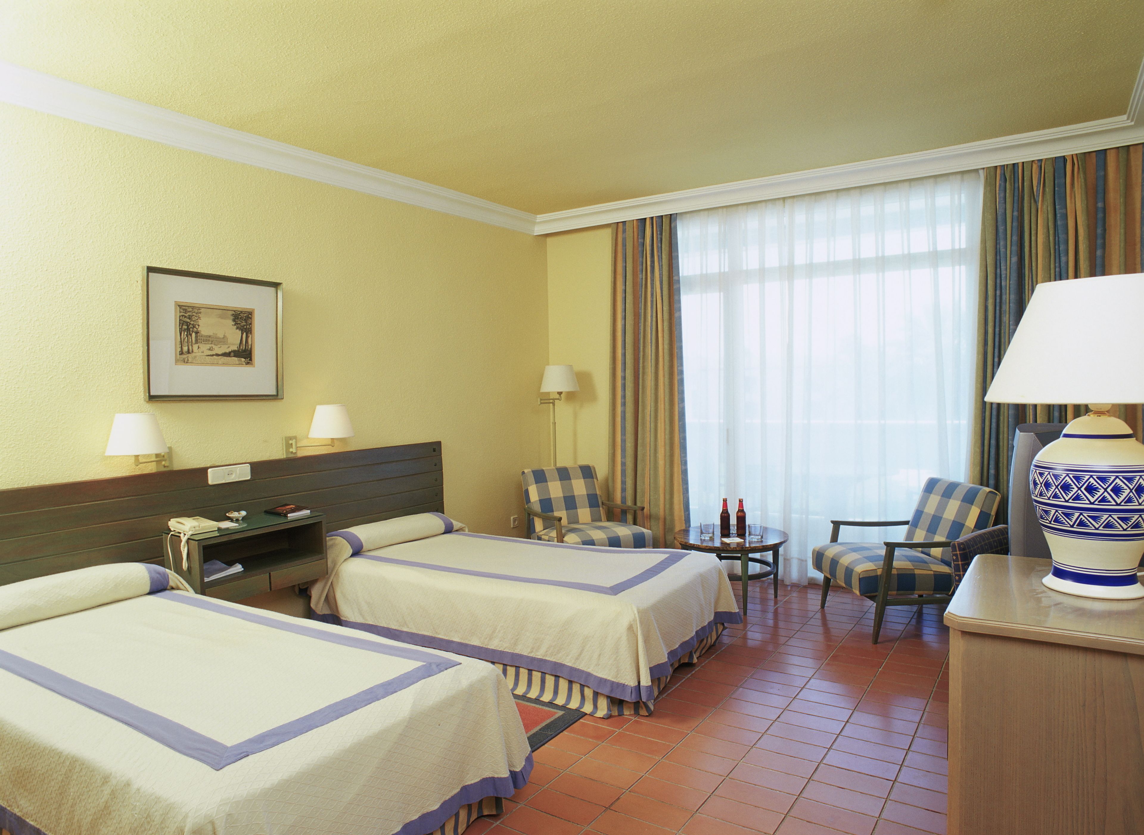 A hotel room with two beds, a desk, a chair, a lamp, and a window with curtains.
