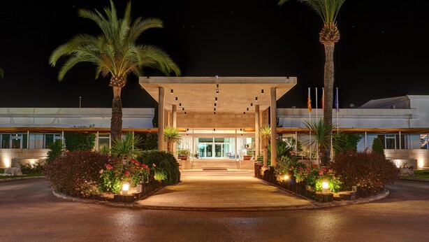 Una entrada de hotel moderna con un paseo techado, palmeras y jardines florales bien iluminados.