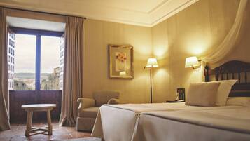 Quarto de hotel com cama, cadeira, mesinha e janela com cortinas.