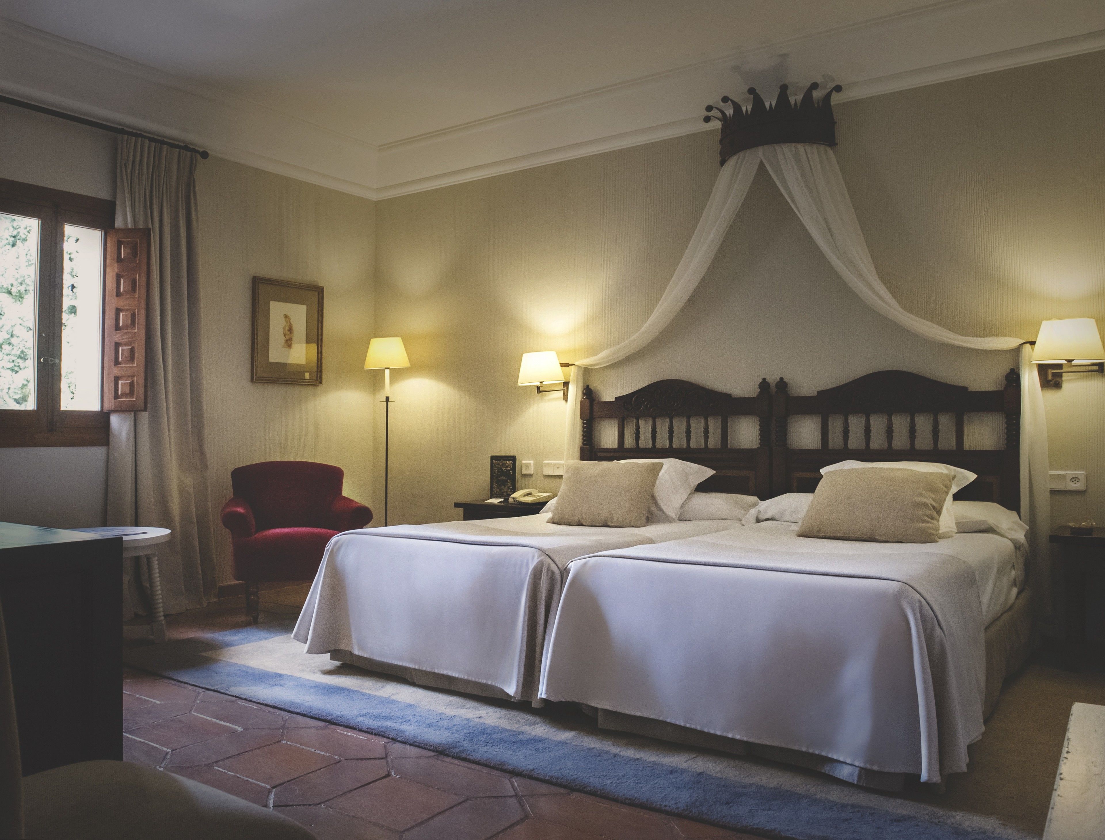 Quarto de hotel com duas camas, uma poltrona vermelha, uma escrivaninha e uma janela com cortinas.