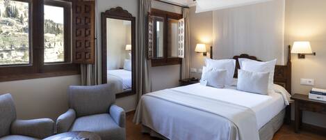 Une chambre d’hôtel avec un grand lit, deux fauteuils, une petite table et une vue sur l’extérieur par une fenêtre.