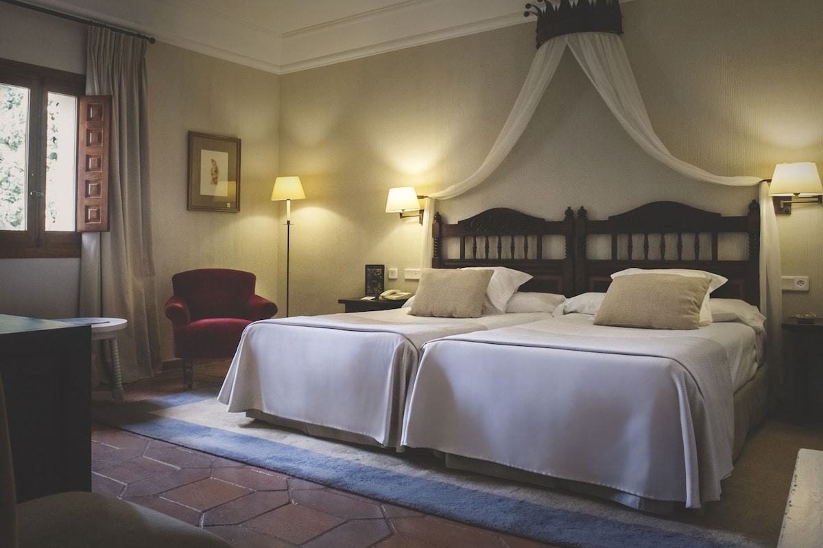 A hotel room with two beds, a red chair, a desk, and a window with curtains.