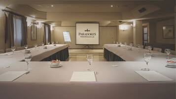 Un salón de conferencias con una larga mesa dispuesta para una reunión, una pantalla de proyector mostrando 'PARADORES Hotels & Restaurants' y un amplio ventanal con cortinas.
