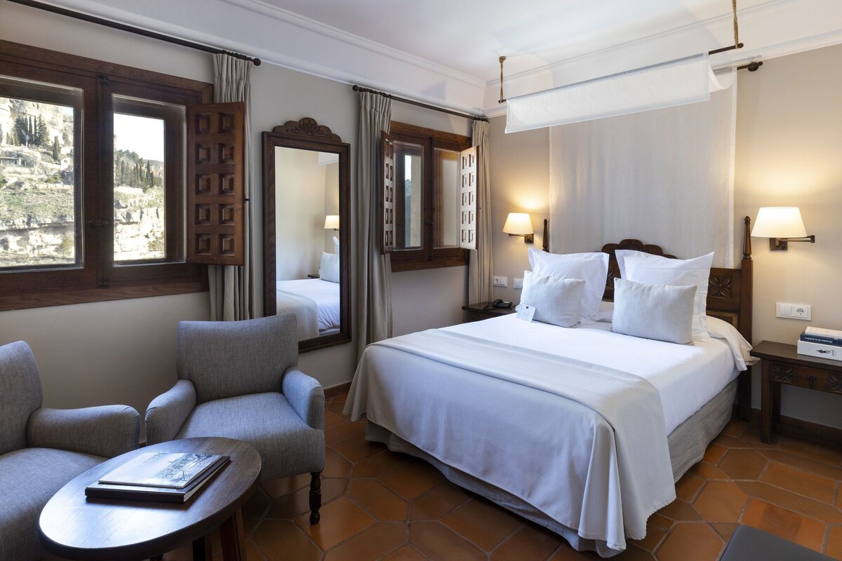 A hotel room with a large bed, two armchairs, a small table, and a view of the outdoors.