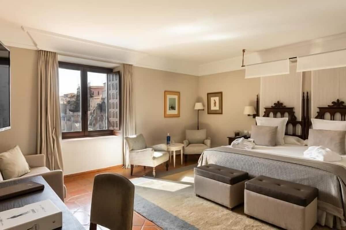 A hotel room with a bed, two chairs, a desk, a small table, and a view of buildings through the window.