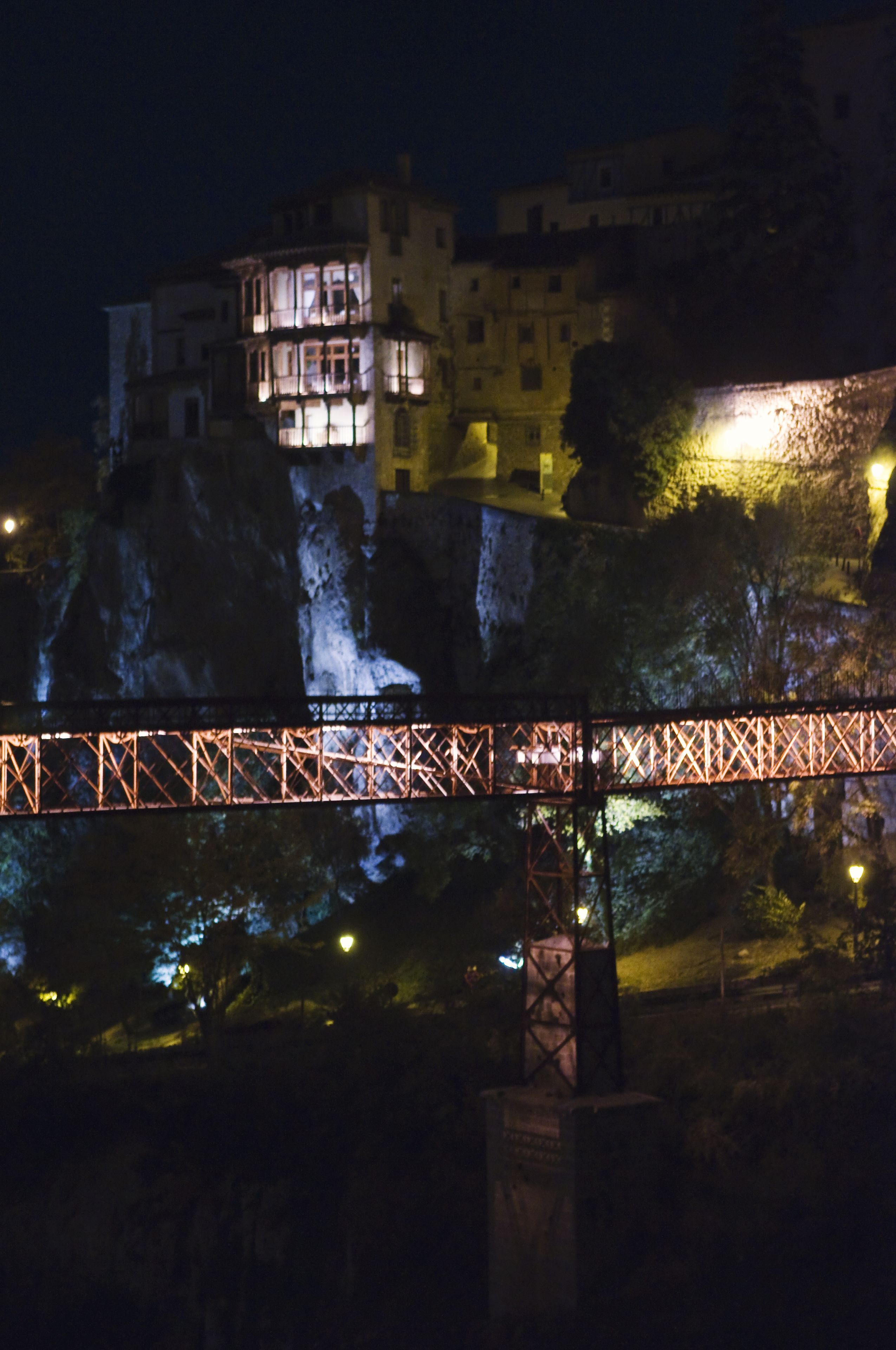 A nighttime view of a bridge with a lit structure and buildings in the background.