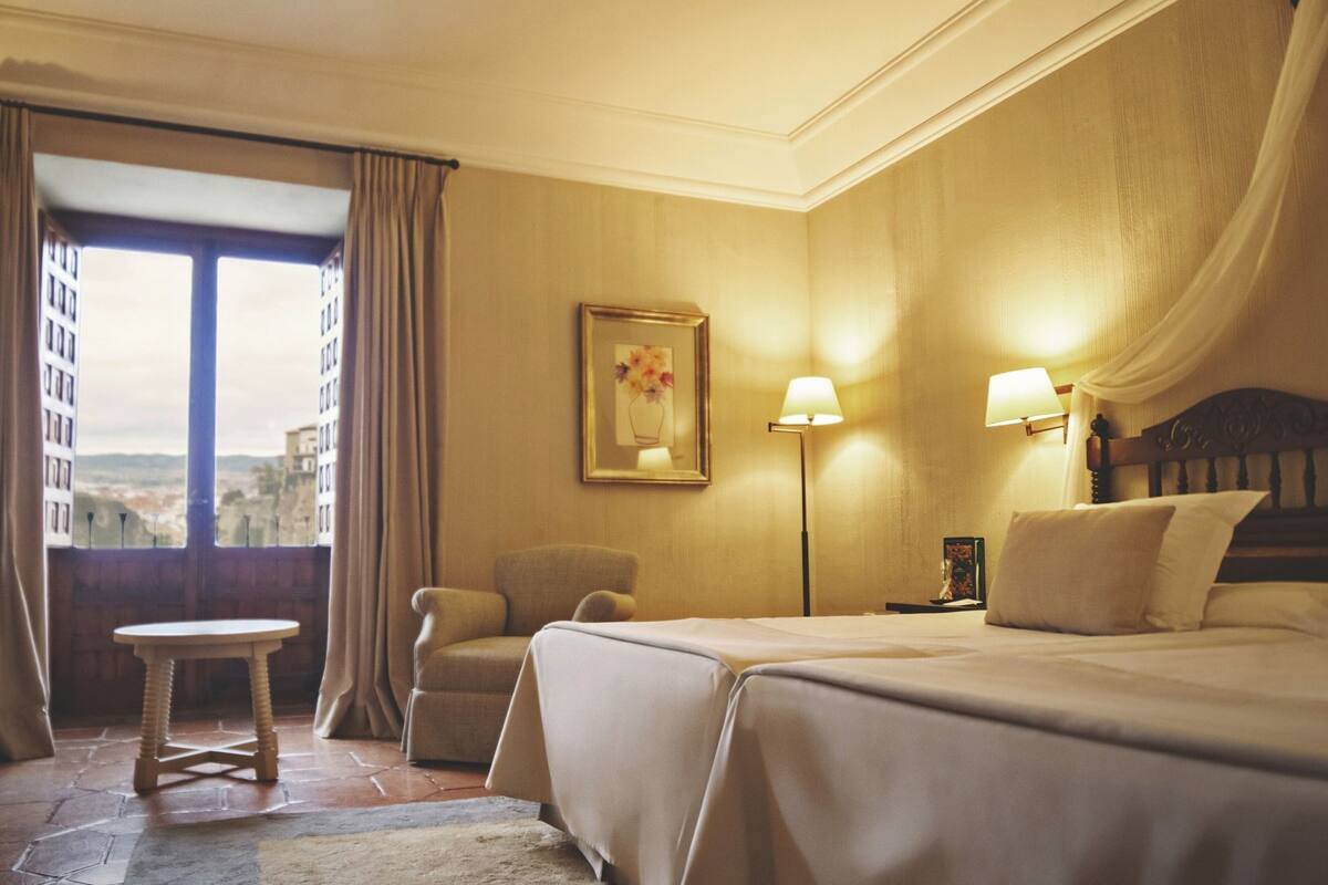 A hotel room with a bed, a chair, a table, a lamp, and a painting.