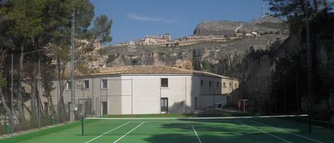 Una cancha de tenis con red, rodeada de árboles y edificios al fondo.