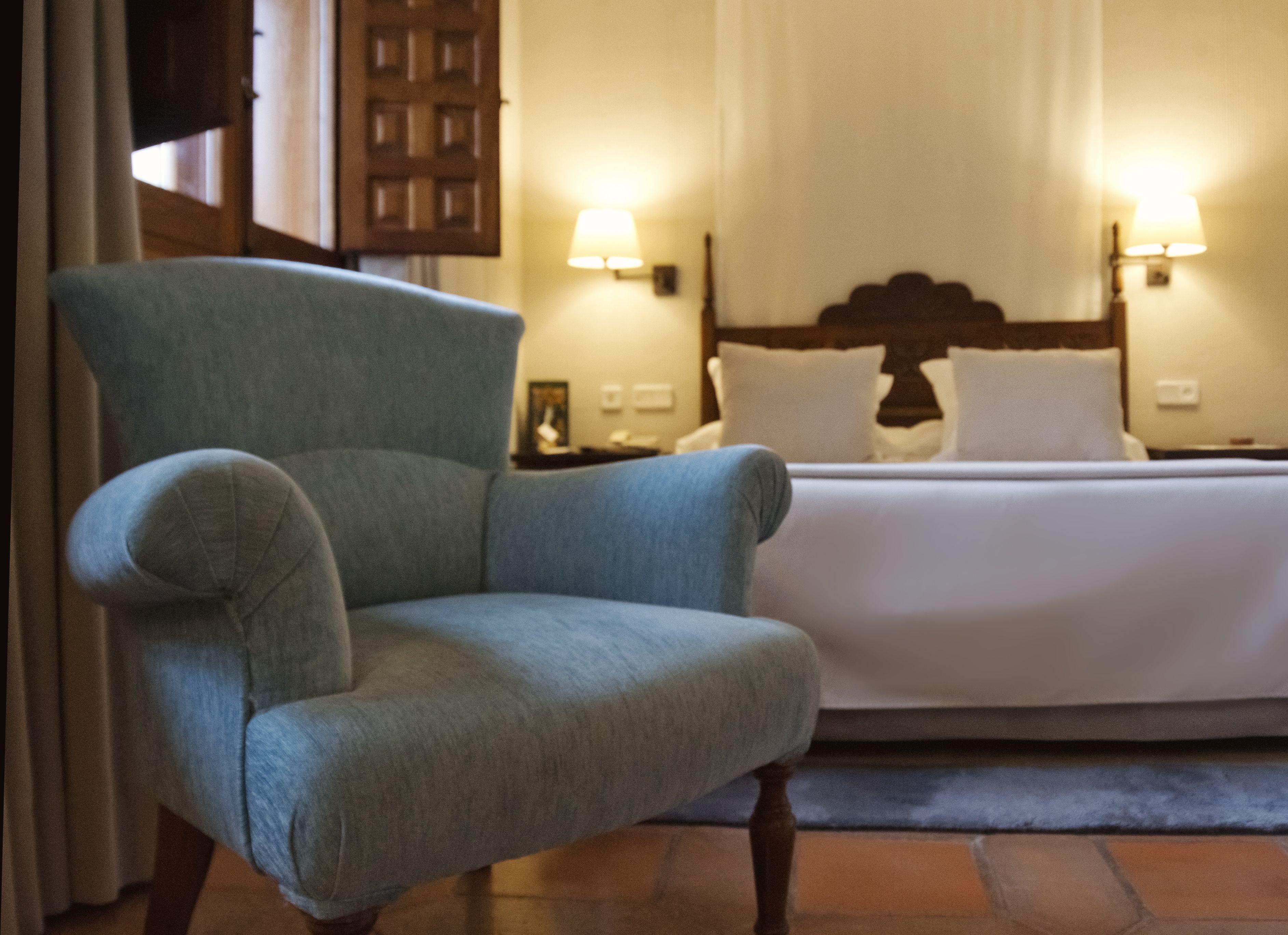 Una habitación de hotel con un sillón azul grande, una cama con ropa de cama blanca, dos lámparas de noche y un cabecero de madera.