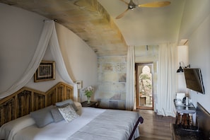A bedroom with a bed, a ceiling fan, a stone wall, a TV, and a view of the outside.
