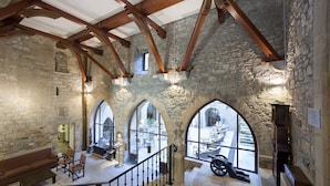 Une pièce aux murs de pierre, avec des poutres en bois, un escalier et des fenêtres en arc donnant sur une cour.
