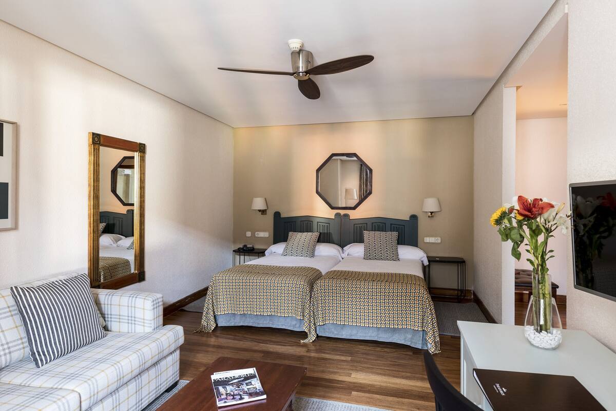 A hotel room with a bed, a sofa, a coffee table, a desk, a mirror, a ceiling fan, and a vase with flowers.