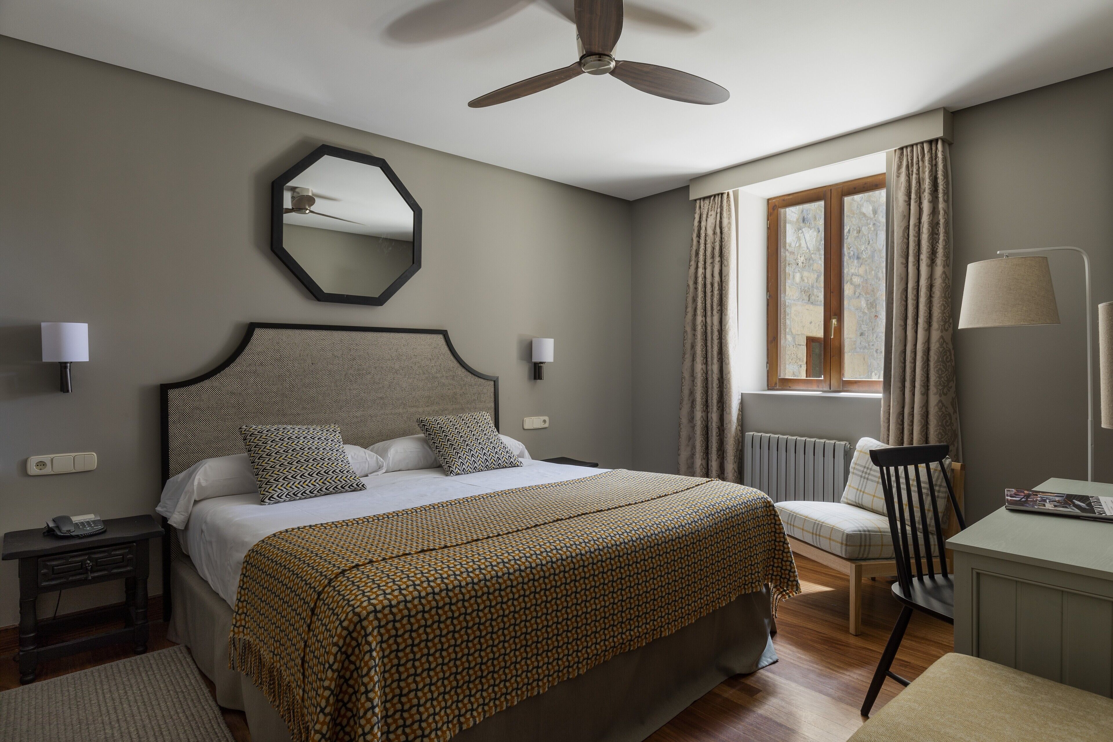 Habitación de hotel ordenada con una cama grande, un ventilador de techo, una ventana con cortinas, una mesita de noche con lámpara y una silla.