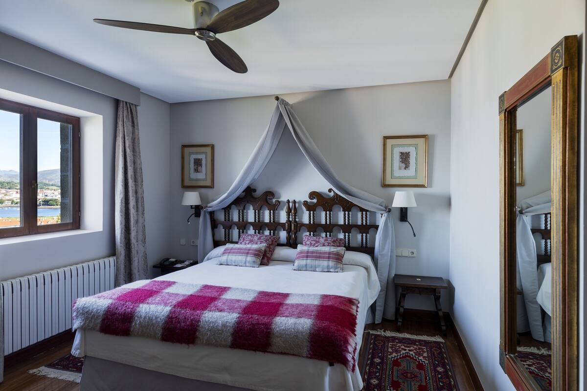 A bedroom with a bed, a ceiling fan, a window with curtains, a bedside table, and a mirror.