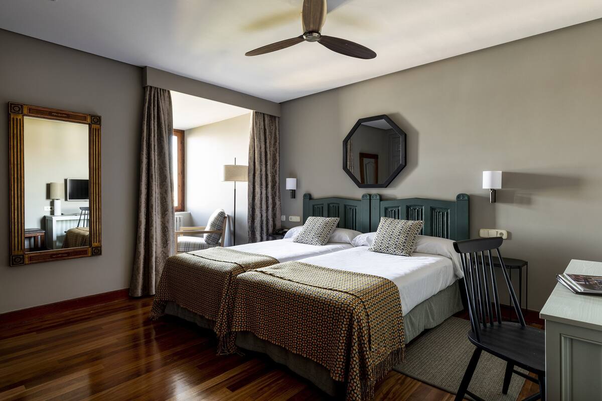 A hotel room with two beds, a ceiling fan, a large mirror, and a view of the outside.