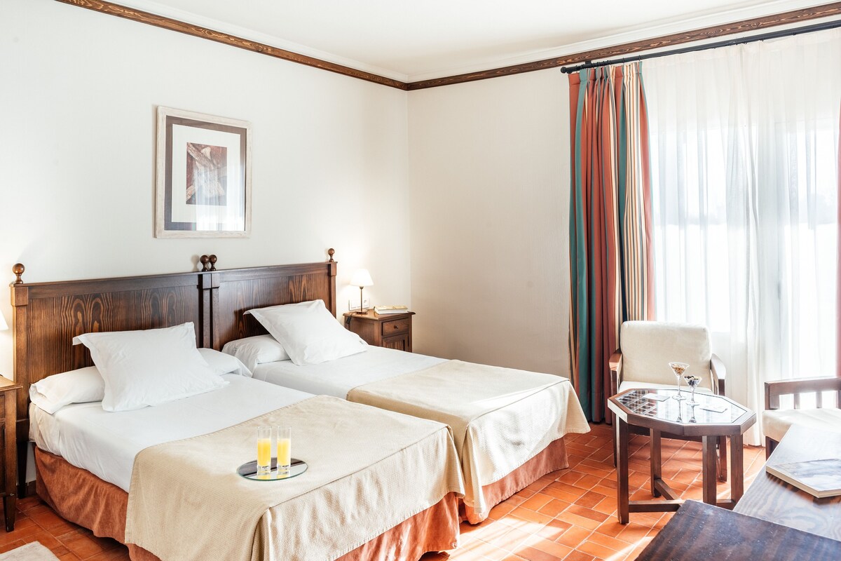 Habitación de hotel con dos camas, una mesita con un vaso de jugo de naranja, una silla y vista al exterior a través de una ventana con cortinas.