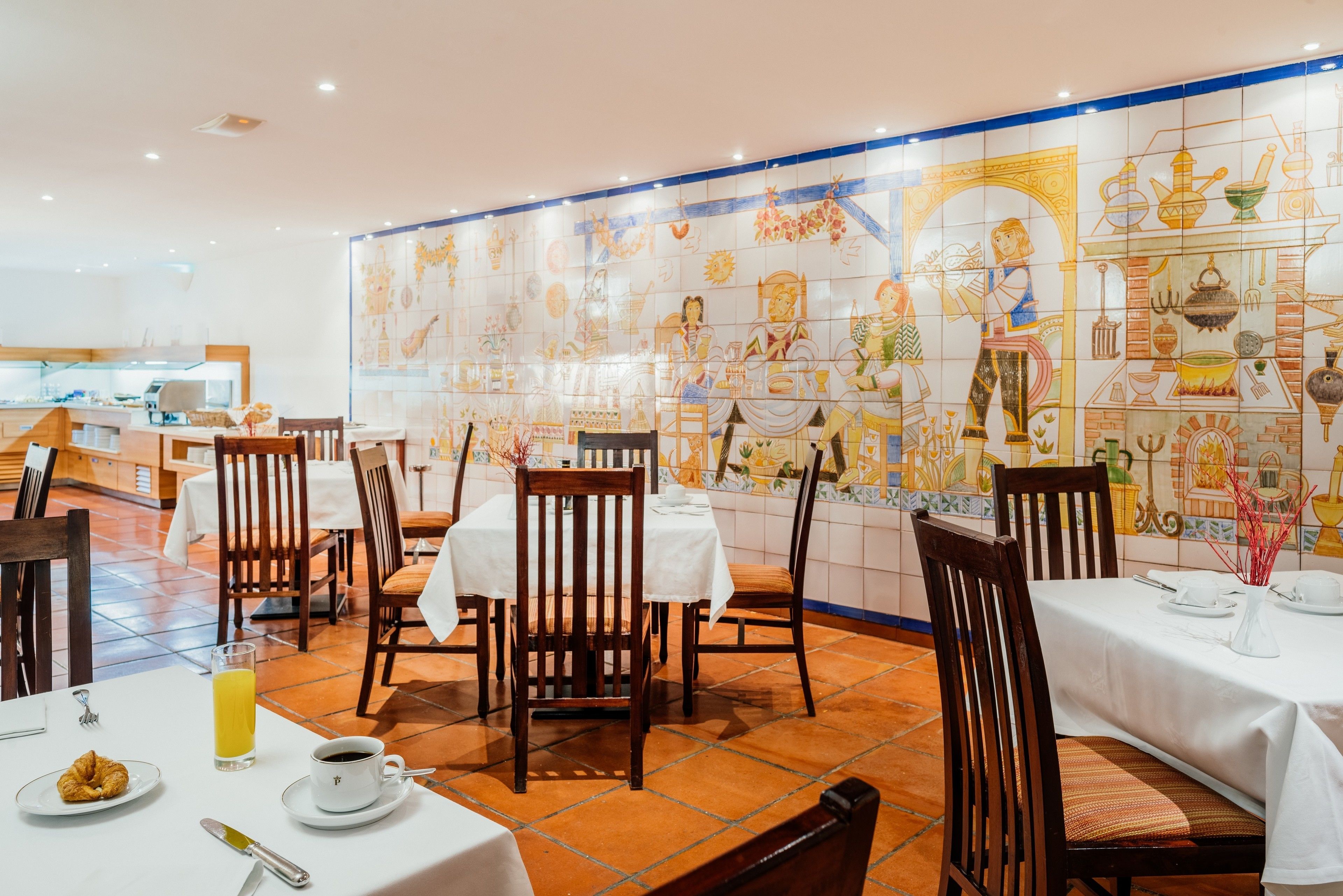 Un restaurante con mesas dispuestas para comer, sillas y una pared de azulejos con ilustraciones.