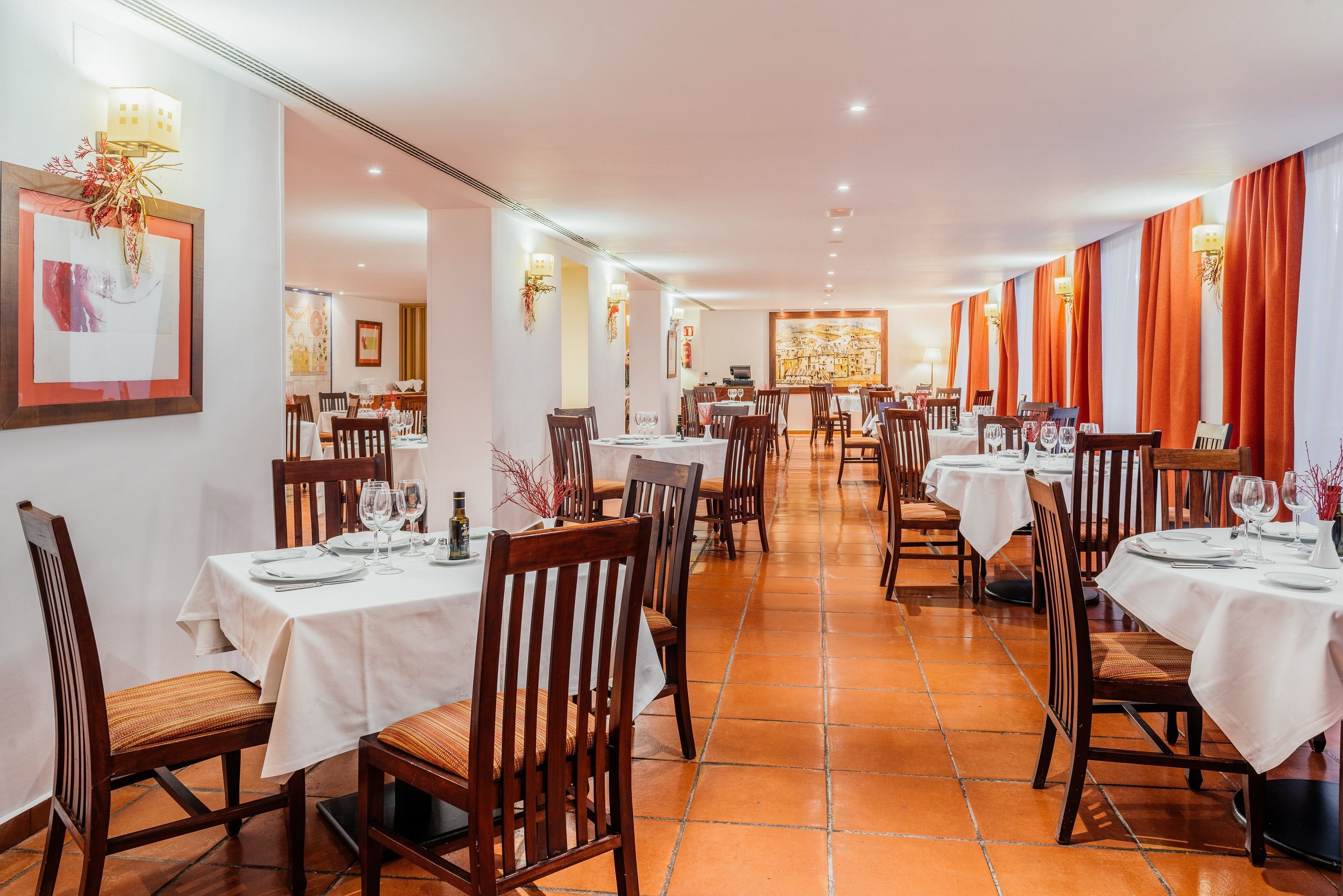 A restaurant with multiple tables and chairs, white tablecloths, and red curtains.