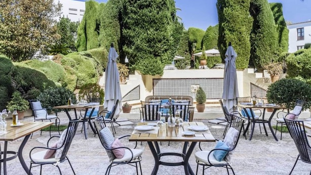 An outdoor patio with tables and chairs, surrounded by neatly trimmed hedges and trees.