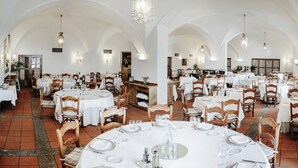 A spacious dining area with multiple tables set for guests, featuring white tablecloths and wooden chairs.