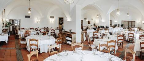 A spacious dining area with multiple tables set for guests, featuring white tablecloths and wooden chairs.