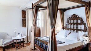 A four-poster bed with a wooden headboard, a bedside table with a lamp, and a chair in a room with a tiled floor. - Parador de Mérida (Merida)