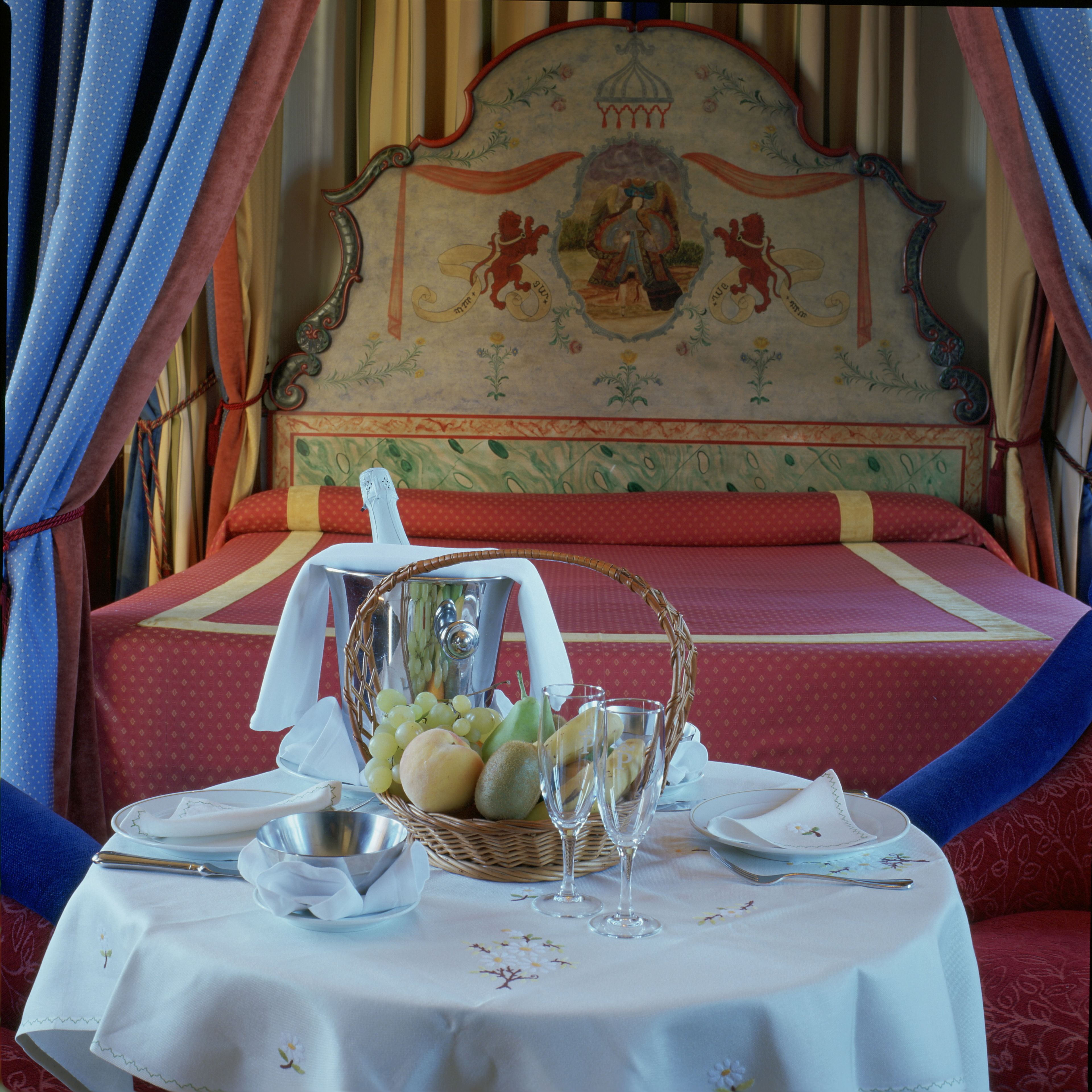 A neatly arranged breakfast table with a basket of fruit, a pitcher, and glassware on a bed with a decorative headboard.