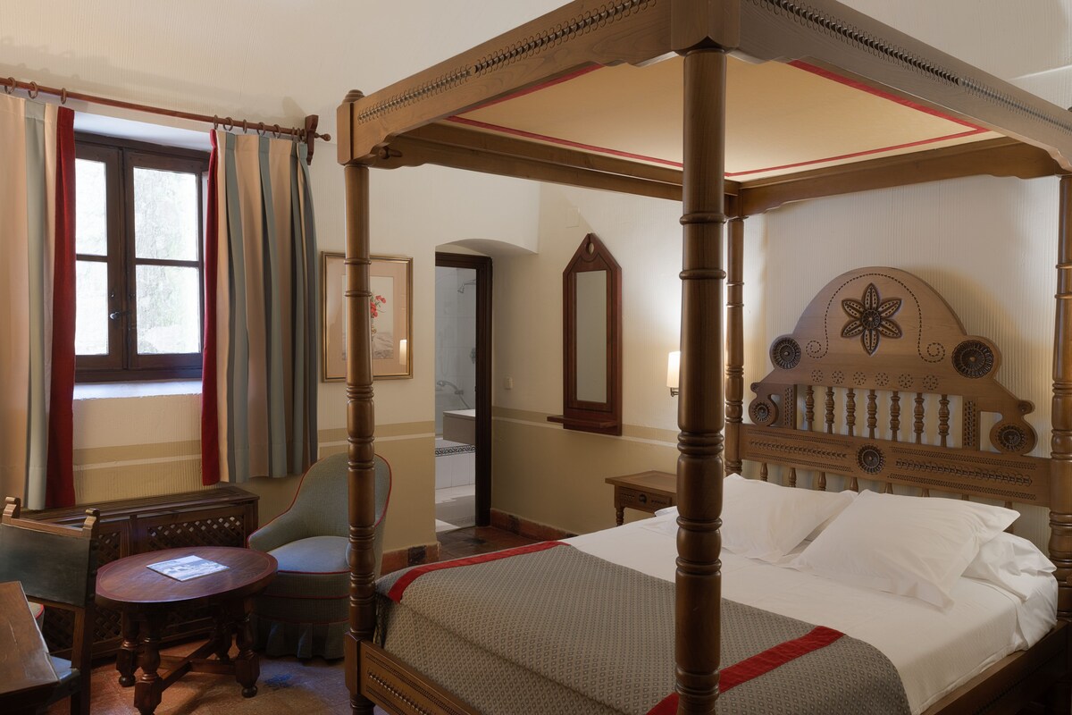 Un lit à baldaquin avec une tête de lit en bois, une table de chevet, une chaise et une fenêtre avec des rideaux.
