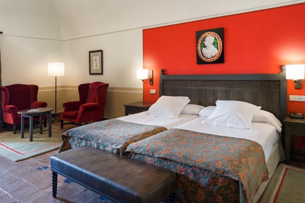 Habitación de hotel con una cama grande, dos sillones rojos, una mesita y un banco.