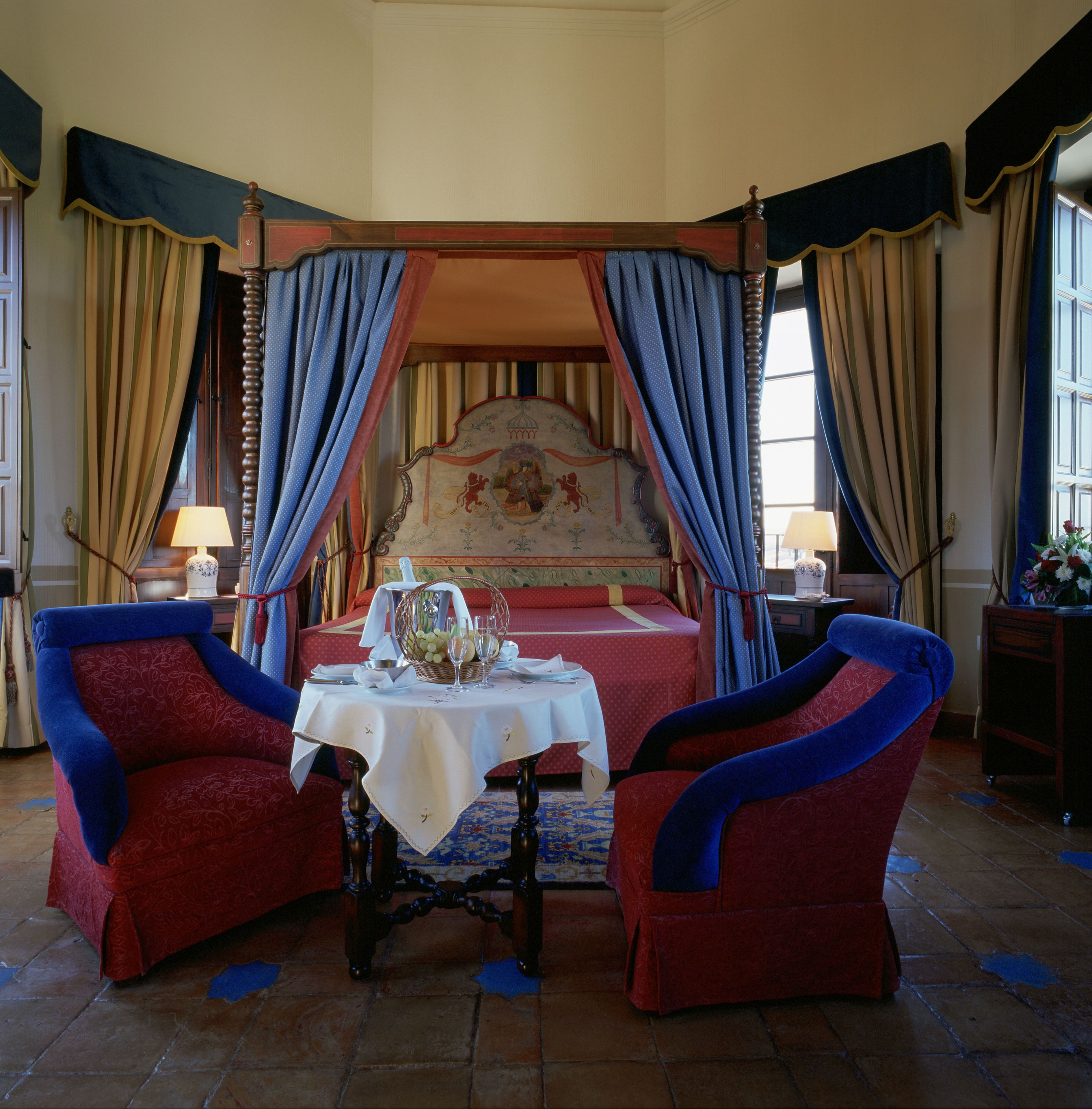 Een hemelbed met een bloemrijk hoofdeinde, een tafel met een vaas en kaarsen, en twee fauteuils.