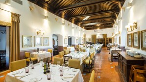 Un vaste espace dédié à la salle à manger, avec plusieurs tables dressées pour les repas, des poutres en bois au plafond et des luminaires fixés aux murs.