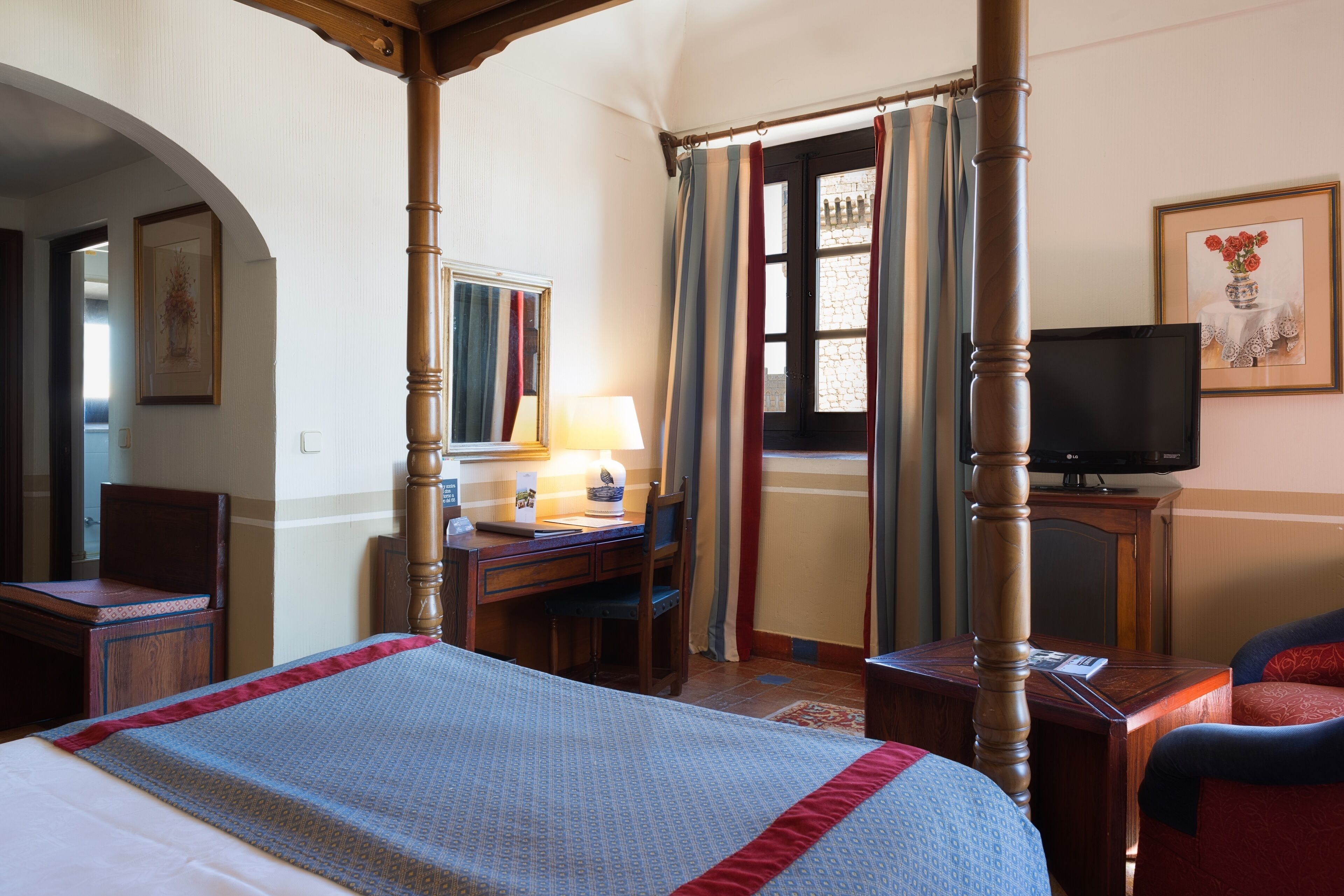 Habitación de hotel con cama con dosel, un escritorio con lámpara, televisor y una silla.