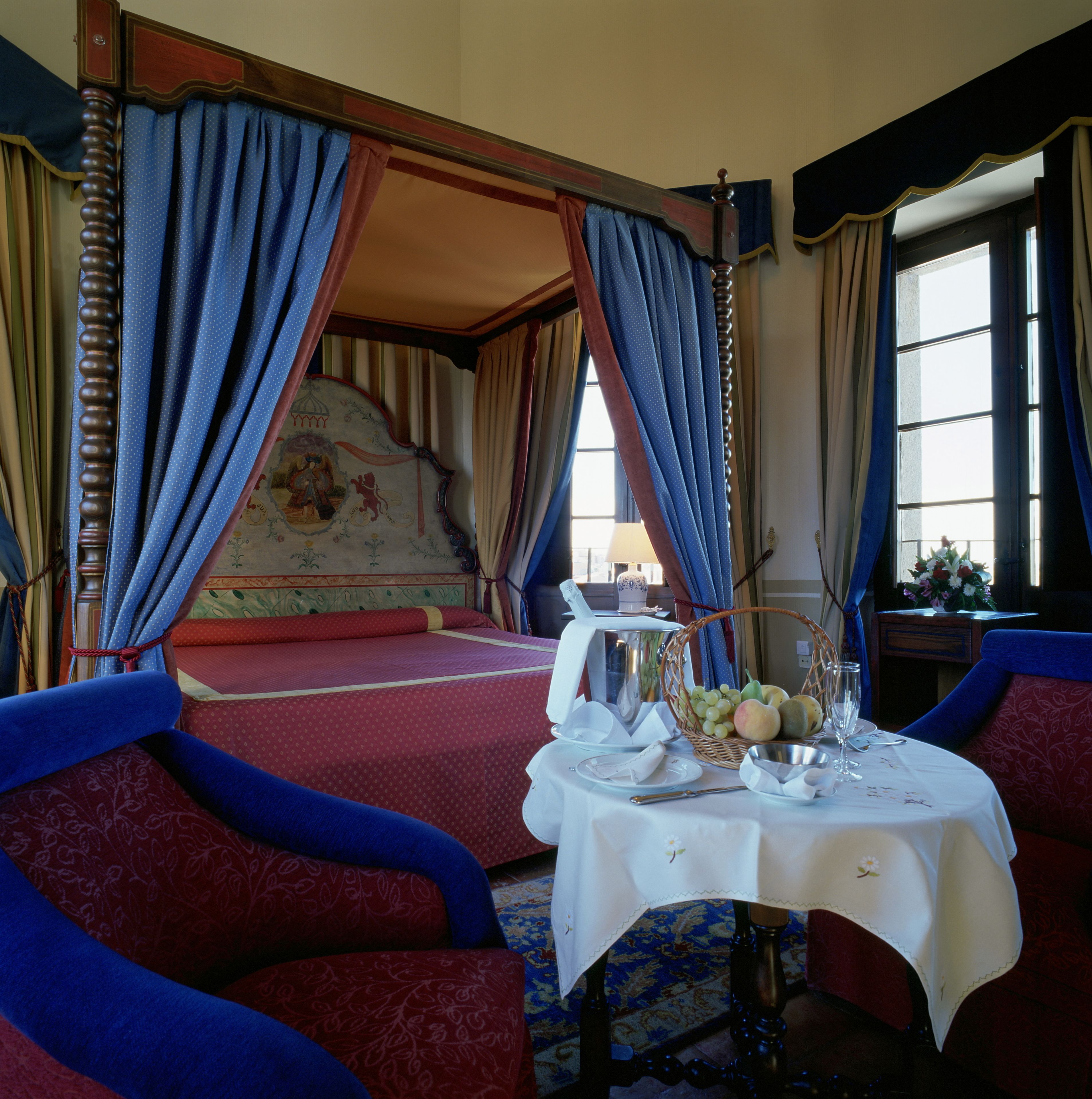 A bedroom with a four-poster bed, a dining table with a fruit basket, and a view of the outdoors through a window.
