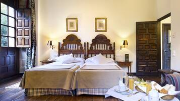 Una cama doble con cabecera de madera, dos mesitas de noche con lámparas, una mesa de desayuno con una cesta de pan y fruta, y una puerta con vista a una ventana.