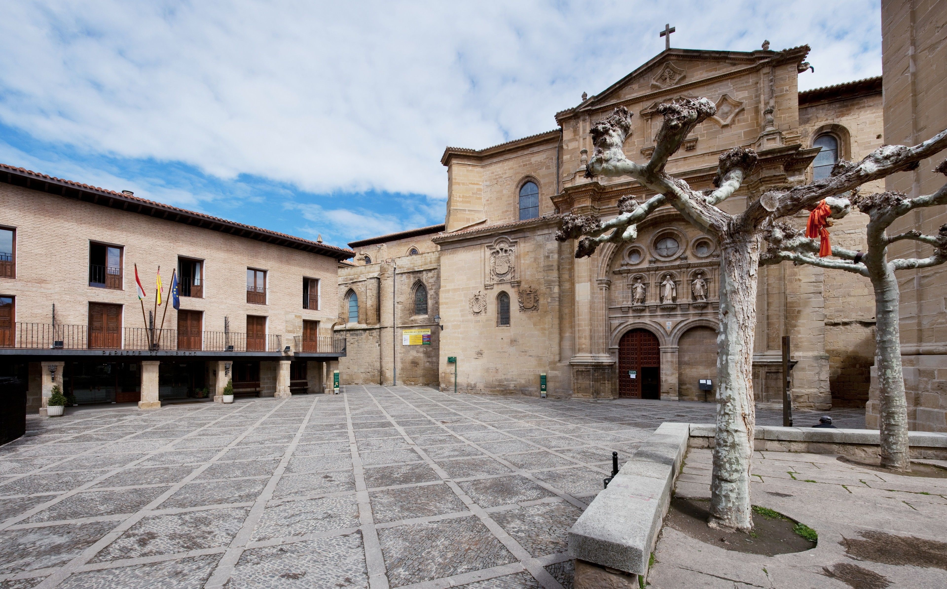 A courtyard with a tiled floor, a building with a cross on top, and a tree with a statue.