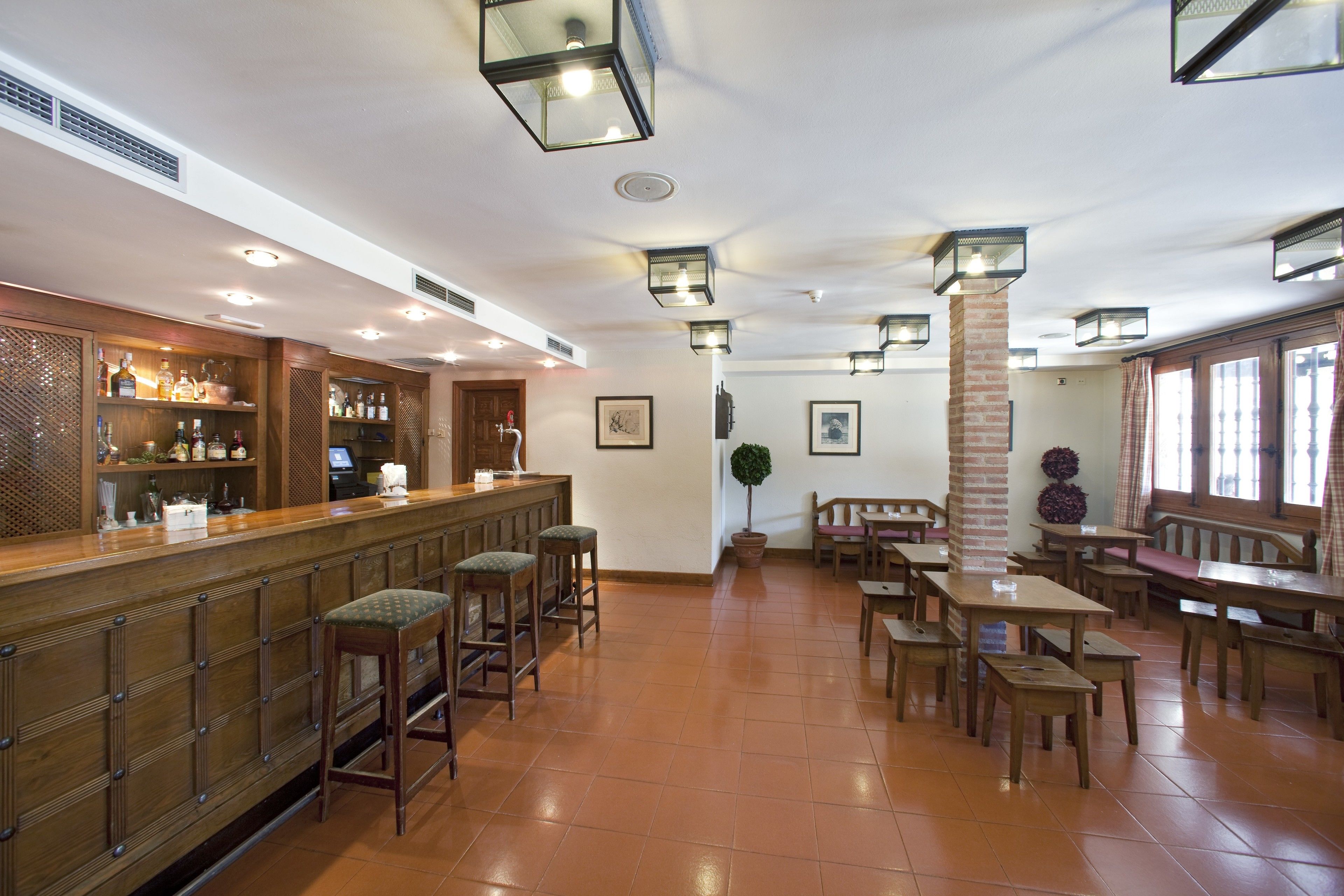 A spacious bar area with a long counter, multiple stools, and a variety of bottles and glasses. There are several tables and benches arranged in the room, with a potted plant and a brick pillar.