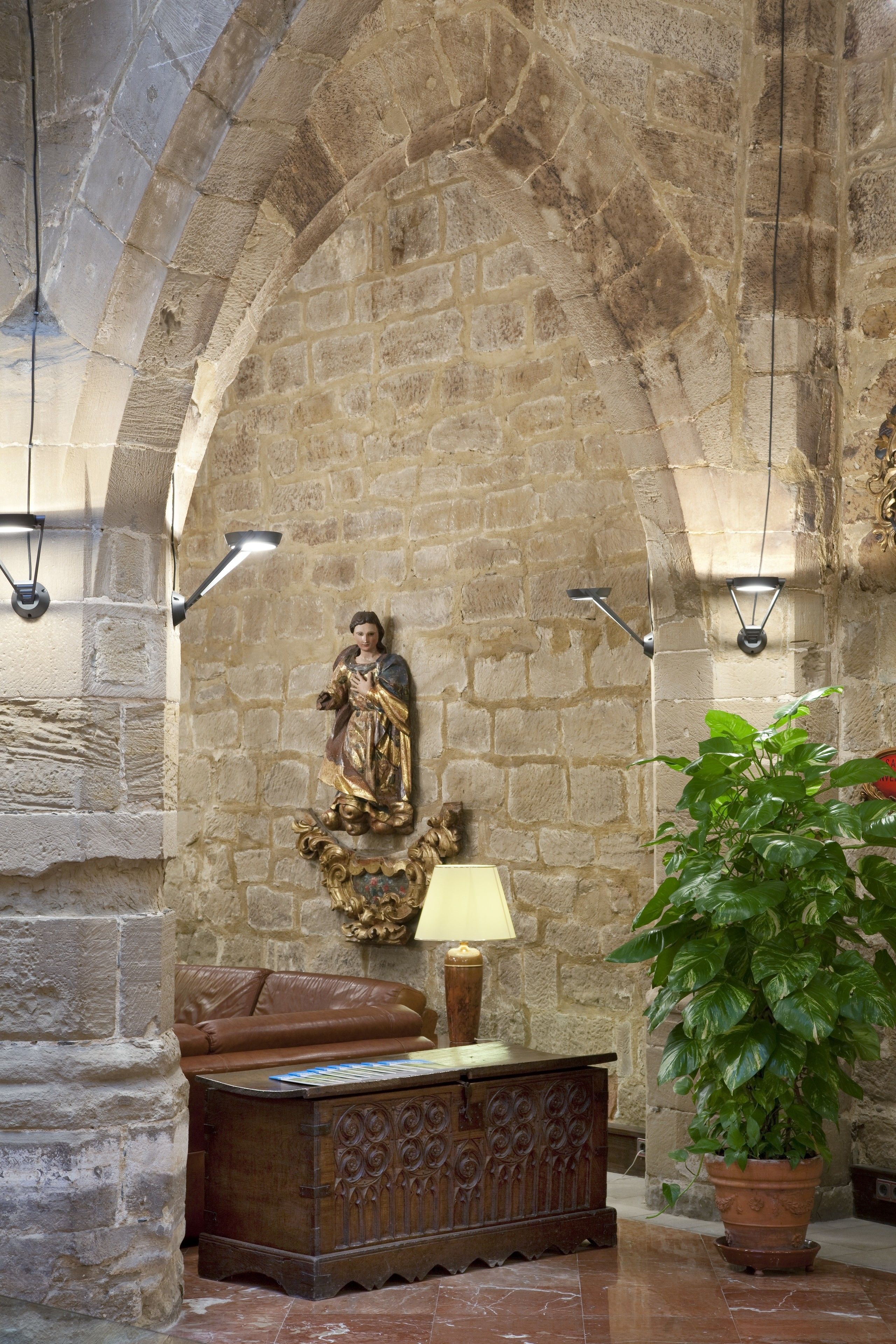 Interior de paredes de piedra con una estatua, un armario de madera, una lámpara y una planta en maceta.