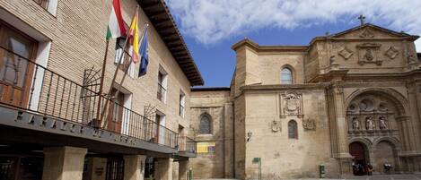 A courtyard with a building featuring a balcony and flags, and a church with an arched entrance.