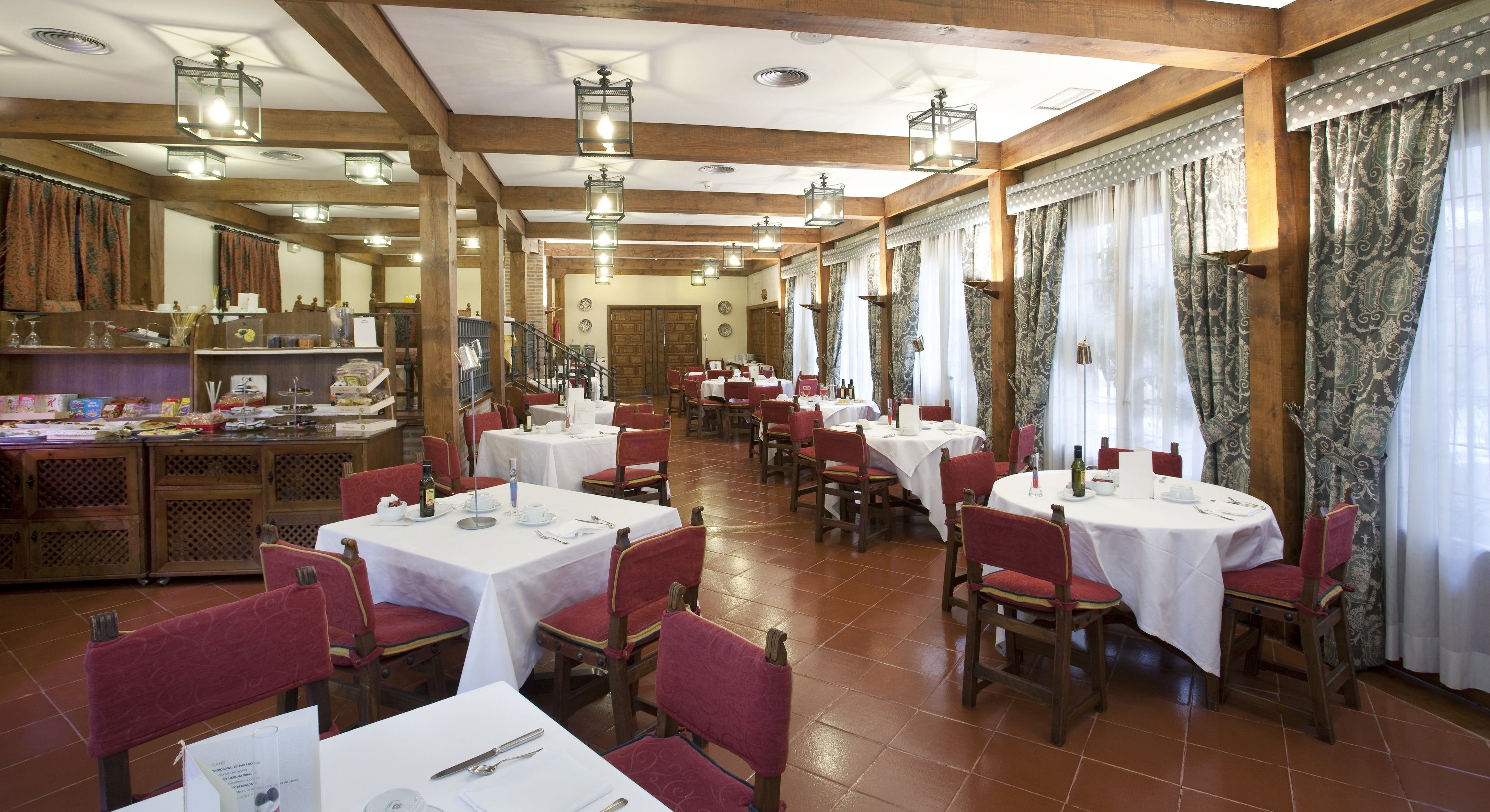 A restaurant with multiple round tables and red upholstered chairs, wooden beams on the ceiling, and patterned curtains.