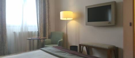 Habitación de hotel con cama, una silla, una mesita, una lámpara y televisor instalado en la pared.