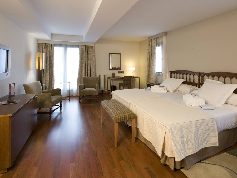 Habitación de hotel con una cama grande, un escritorio, una silla, un banco, un televisor y una ventana con cortinas.
