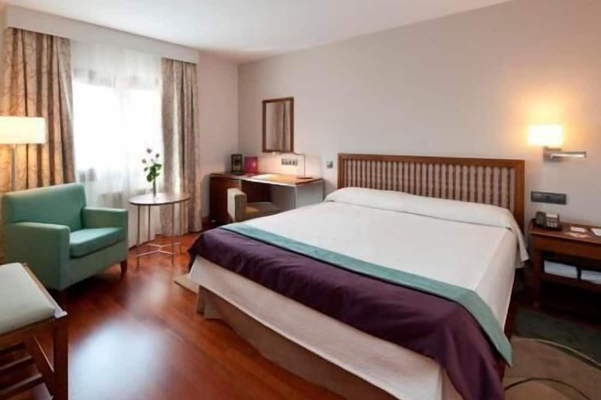 A hotel room with a bed, a desk, a chair, and a lamp.
