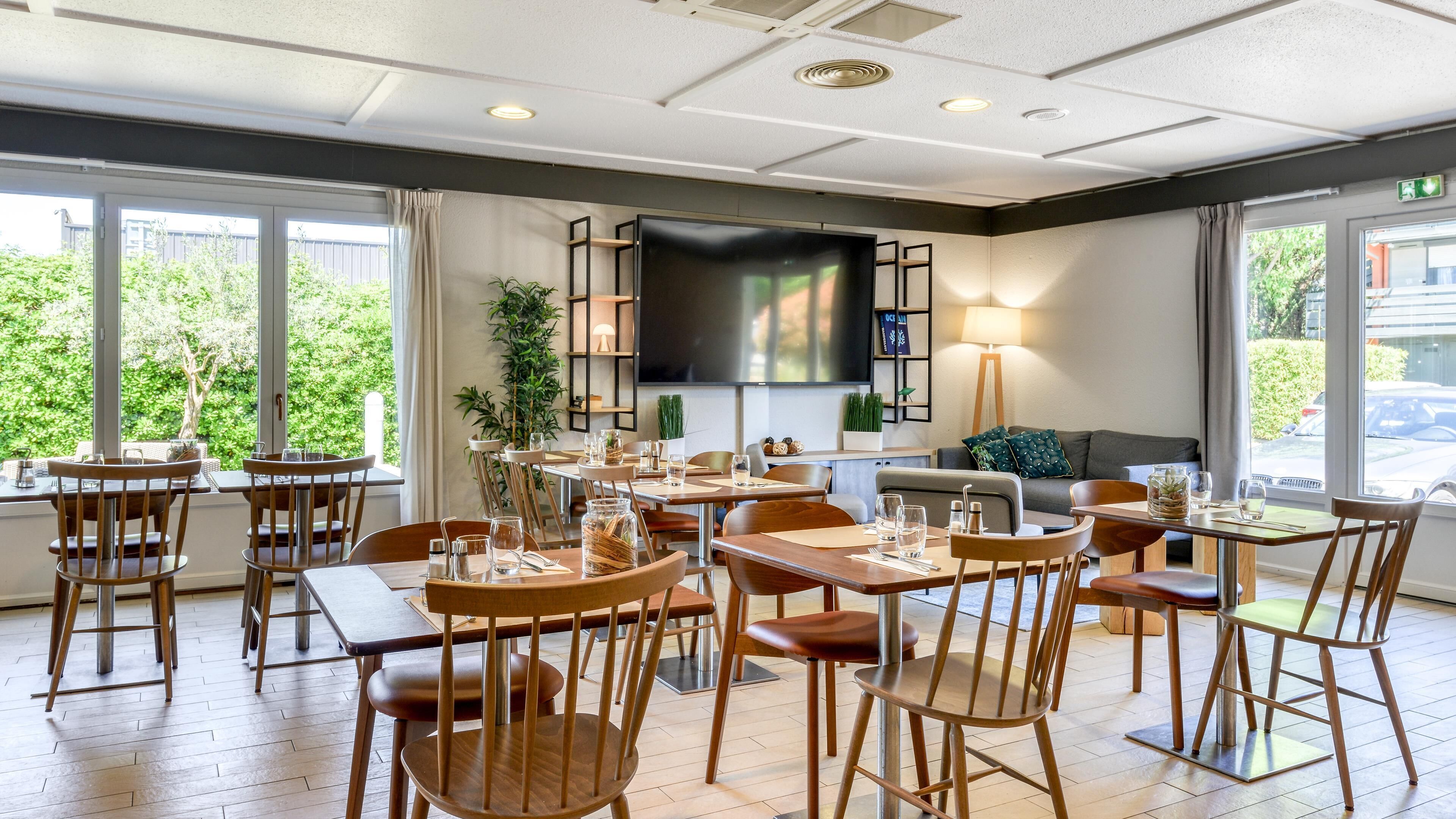 A modern dining area with wooden tables and chairs, a large flat-screen TV, and a sofa in the background.
