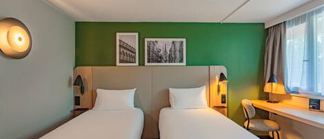 Une chambre d’hôtel avec deux lits, un bureau, une chaise, une fenêtre avec des rideaux et deux tableaux encadrés au mur.