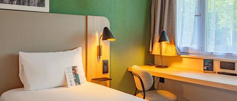 Habitación de hotel con cama, escritorio con silla, lámpara, ventana con cortinas y panel de control en la pared.
