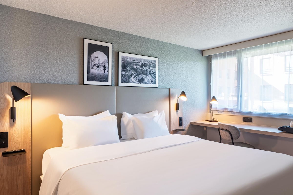A hotel room with a bed, two wall-mounted picture frames, a desk with a lamp, and a window with curtains.