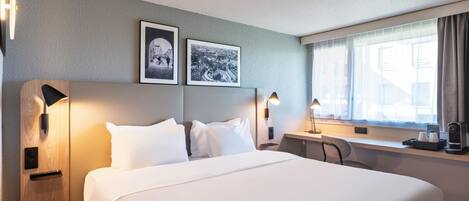 Habitación de hotel con una cama, dos cuadros colgados en la pared, un escritorio con lámpara y una ventana con cortinas.