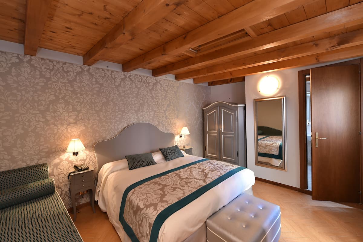 A bedroom with a wooden ceiling, a bed with a patterned bedspread, a nightstand with a lamp, a wardrobe, and a mirror on the wall.