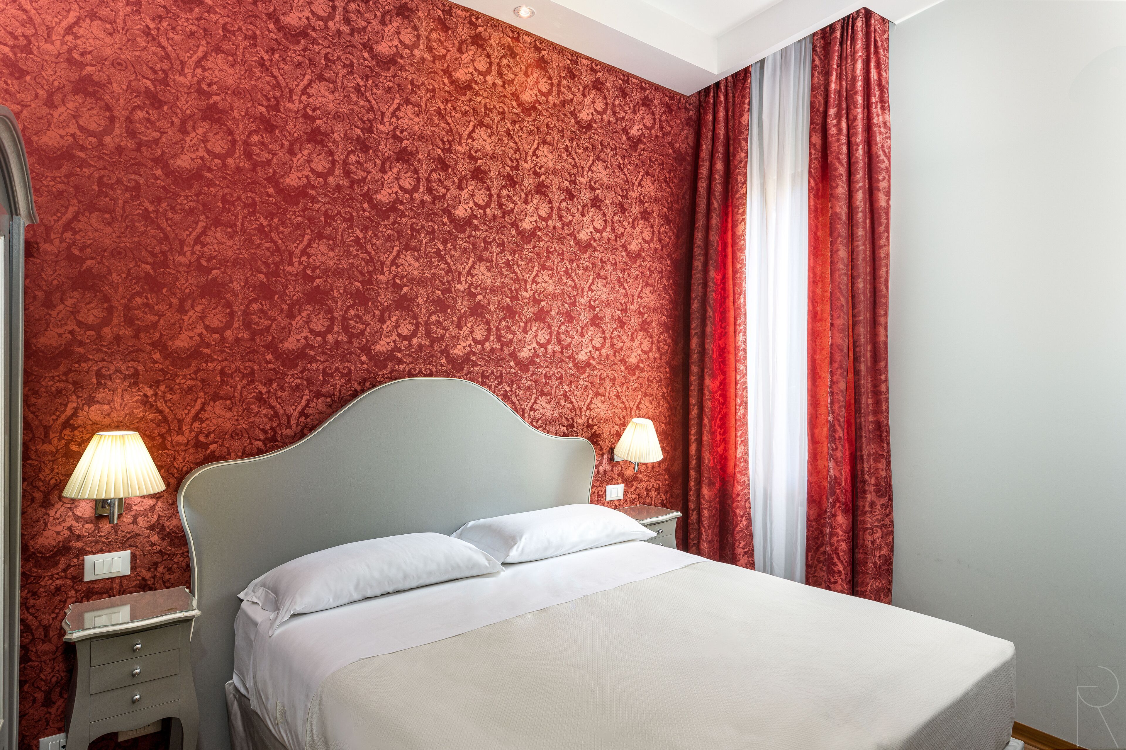 Una camera d'albergo con una parete rossa decorata, un letto con lenzuola bianche, due lampade sui comodini e una finestra con tende rosse.