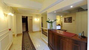 Zona de recepción de hotel con un mostrador de madera, un jarrón con flores, un cuadro en la pared y un pasillo con alfombra que conduce a una escalera.